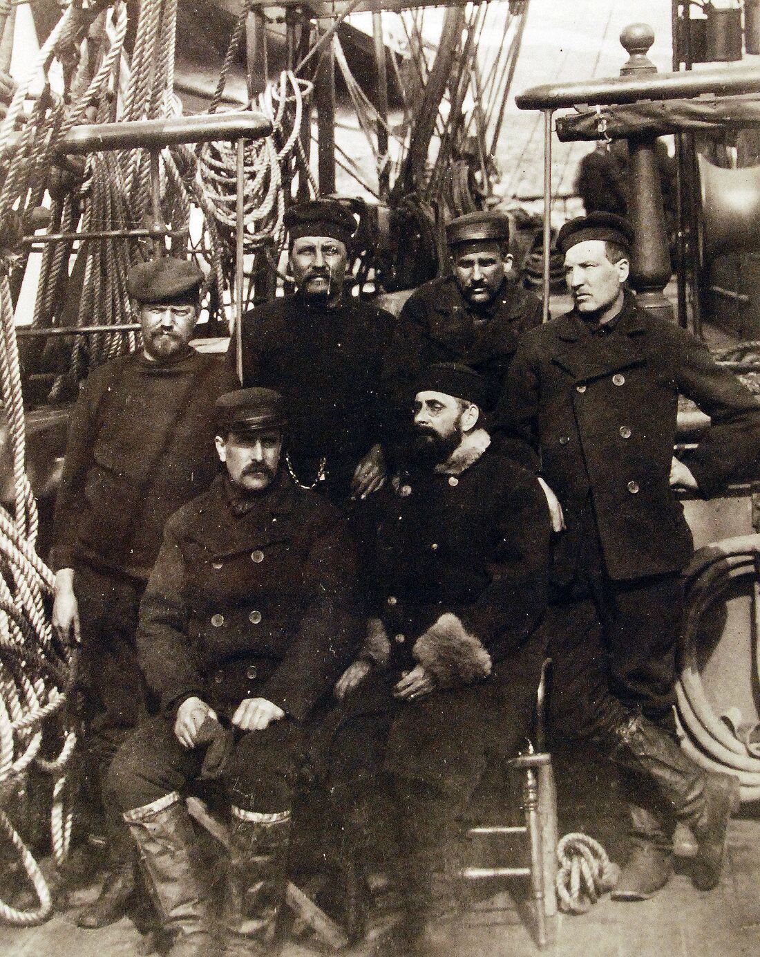 Sepia photograph of six gaunt, heavily bundled expedition survivors posed among the rigging on the deck of the rescue ship USS Thetis in 1884.